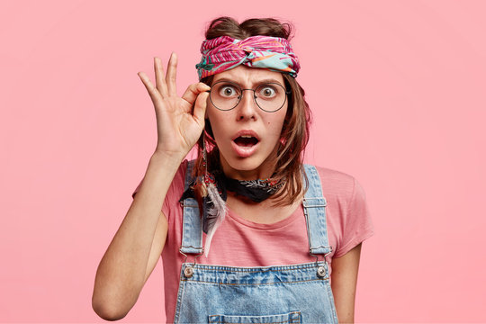 Scared surprised beautiful female hippy stares through glasses, can`t believe in bad news, dressed in overalls and headband, isolated over pink studio wall. People, emotions, reaction concept