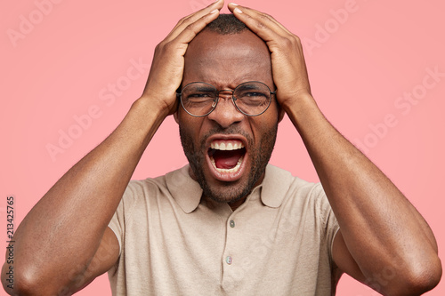 Fotografía Close up portrait of desperate African American male regrets something wrong, frowns face in discontent, opens mouth widely, dressed in t shirt, isolated over pink background