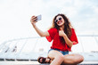 © Maksym Azovtsev - Cute curly girl in sunglasses takes a selfie on smartphone, showing a thumb up sitting on marble slab outdoors. Dressed in stylish clothes.