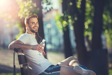 Full Length Portrait Of Delighted Athlete Sitting On Bench Outdoors. He Is Drinking Water And Listening To Music With Joy
