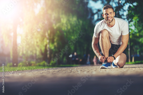 Full length portrait of smiling athlete kneeling and fixing shoelaces outdoors Poster Mural XXL
