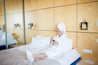 © pressmaster - Young restful female in white bathrobe, towel on head and flippers sitting on bed and texting in smartphone