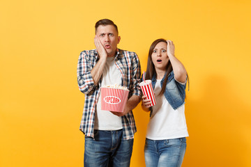  Young shocked amazed couple woman man watching movie film on date holding bucket of popcorn and plastic cup of soda or cola clinging to head isolated on yellow background. Emotions in cinema concept.