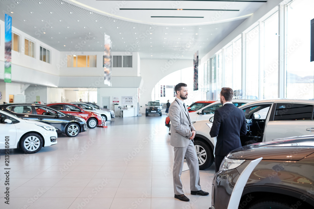 Professional salesperson selling new cars in a modern auto salon. Two men standing and choosing a new car - obrazy, fototapety, plakaty 