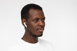© Damir Khabirov - Horizontal headshot of young African American man pictured isolated on grey background with short hair and white wireless earphones listening to audio tracks or conversation with concentrated face