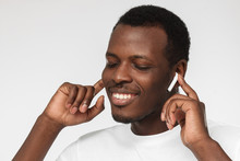 Headshot Photo Of Young Handsome African Man Turned Leftwards Against Grey Background Absorbed With Good Sound Quality Of New Wireless Earphones, Eyes Shut With Fascination And Satisfaction
