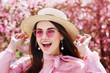 © Victoria Fox - Outdoor close up portrait of young beautiful happy smiling woman wearing straw hat, pink sunglasses, earrings, blouse, posing street, near blooming tree. Advertisement concept