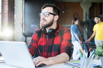 Portrait of modern bearded man wearing glasses and red shirt using laptop looking away pensively in sunlit office of IT developers team, copy space