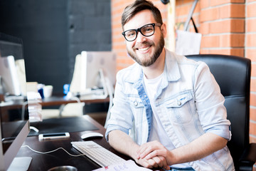 Portrait of contemporary business manager wearing casual clothes smiling happily at camera while sitting at computer desk in modern office