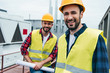 © LIGHTFIELD STUDIOS - smiling engineers in safety vests and helmets with blueprint on roof