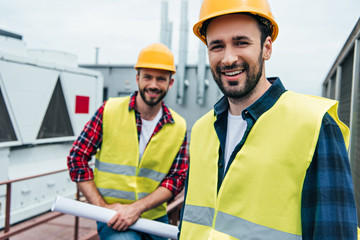 smiling engineers in safety vests and helmets with blueprint on roof