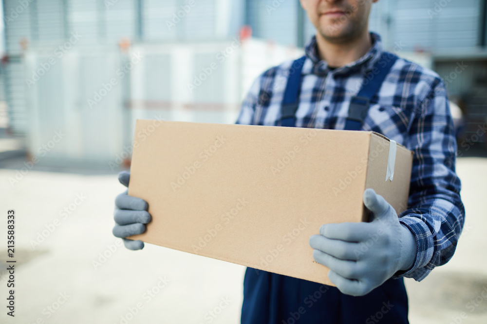 Close-up of moving company worker in gloves holding cardboard box and carrying it at container storage area - obrazy, fototapety, plakaty 