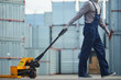 Unrecognizable worker in overall carrying load cart and walking over cargo container area