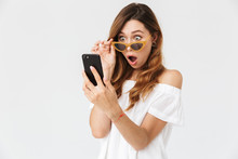 Portrait Of Surprised Young Woman 20s Looking At Smartphone From Under Sunglasses With Open Mouth, Isolated Over White Background