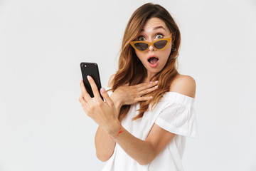 Canvas Print - Portrait of caucasian surprised woman 20s holding smartphone and looking at you from under sunglasses with open mouth, isolated over white background