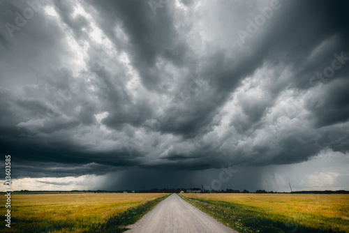 Fotografie, Obraz Gravel road in the middle of green and yellow fields and stormy clouds above it