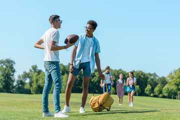 Wall Mural - multiethnic boys playing with rugby ball while classmates walking behind in park