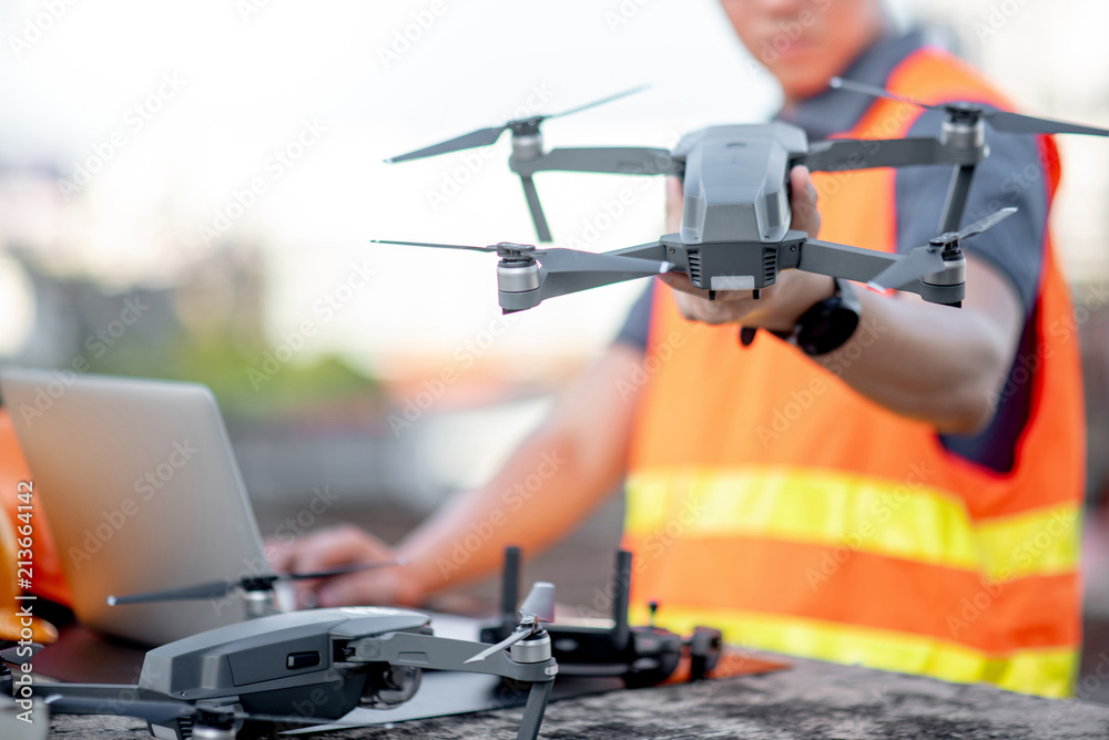 Young Asian engineer man working with drone laptop and smartphone at construction site. Using unmanned aerial vehicle (UAV) for land and building site survey in civil engineering project. - obrazy, fototapety, plakaty 