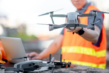 Young Asian Engineer Man Working With Drone Laptop And Smartphone At Construction Site. Using Unmanned Aerial Vehicle (UAV) For Land And Building Site Survey In Civil Engineering Project.