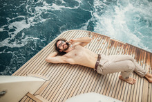 Man Relaxing Under The Sun, Lying On A Wooden Deck Of The Boat At Sea. Luxury Vacation On A Yacht In The Islands.