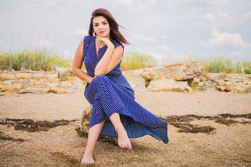 Brunette girl in a long blue dress sitting on the seashore