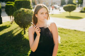 Young attractive girl smoking e-cigarette at the sunny weather outdoors, looking at camera and vaping.