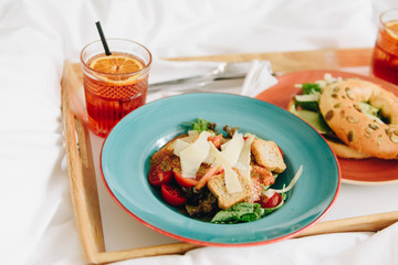 Breakfast or lunch in bed of a panzanella salad with croutons and a bagel with fresh lettuce, two drinks and French fries on a tray in colorful plates