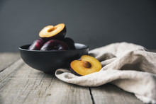 Close Up Of Plums In Bowl With Napkin On Wooden Table Against Gray Background
