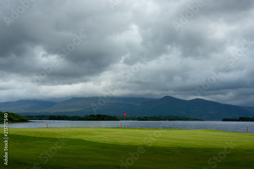 Fotografie, Obraz Dramatic golf course landscape with green tee,red flag and beautiful mountain views across lake