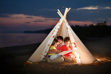 Mom and two children are reading a book with flashlights in tent in the evening on the river bank.