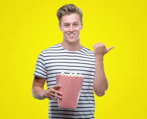 Sticker - Young handsome blond man eating popcorn pointing with hand and finger up with happy face smiling