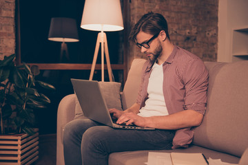 Wall Mural - Portrait of attractive, busy,  smart guy, hard worker holding arms on keypad, looking at screen of laptop on his legs, sitting in living room, working at night, taking work at home