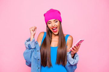 Charming and happy girl joyfully clasps her hand in a fist and holding the phone in her hands isolated on bright pink background