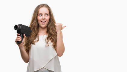 Wall Mural - Young blonde woman holding super 8 video camera pointing and showing with thumb up to the side with happy face smiling