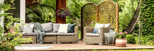 Panorama Of Wicker Garden Furniture With Cozy Pillows And Blankets On A Wooden Terrace In Beautiful Outdoor Greenery