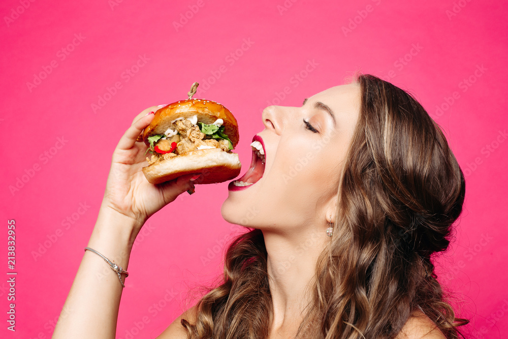 Close up of hungry girl with opened mouth, holding and eating big hamburger. Pretty woman with curly hairstyle and red lips eating tasty cheeseburger with tomato and meal. Concept of fast food.
