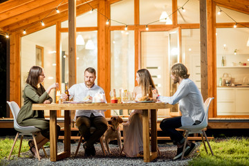 Wall Mural - Friends having a dinner sitting at the table on the backyard of the modern house decorated with lights during the evening time outdoors