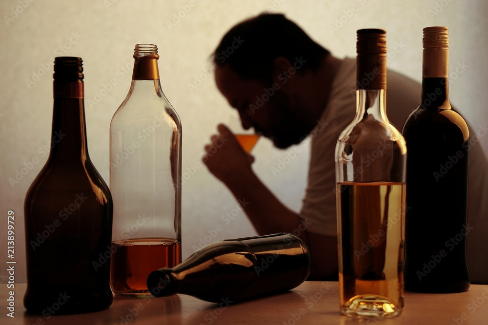 silhouette of anonymous alcoholic person drinking behind bottles of alcohol  - obrazy, fototapety, plakaty 