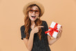 © Drobot Dean - European summer woman 20s wearing straw hat and sunglasses smiling while holding present box with red ribbon, isolated over beige background