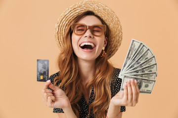 Sticker - Image of happy young woman 20s wearing straw hat and sunglasses laughing and demonstrating credit card and fan of dollar cash, isolated over beige background