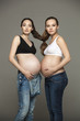 Две привлекательные беременные женщины стоят прислонившись животами друг к другу на сером фоне. Последние месяцы беременности.