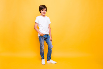 Full height portrait of attractive young cheerful school boy, smiling standing holding hand in jeans pocket over yellow background, isolated. Copy space