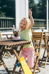 Wall Mural - selective focus of happy child with sticking out tongue doing peace gesture at table with delicious dessert in cafe