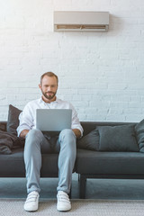 Wall Mural - handsome bearded man using laptop on sofa, with air conditioner on wall behind