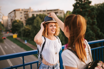 Young woman puts hat on her female friend's head on city street. Summertime fun.