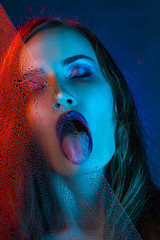 A beautiful model girl, illuminated with blue and red lights, half peeks out of a glass covered with drops of water and licks a water from the glass