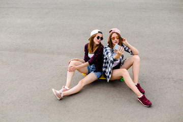 two young happy girl friends in hipster outfit sitting together on longboard and making selfie on ph