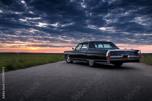 Plakat Czarny retro vintage muscle car jest zaparkowany na wiejskiej drodze asfaltowej na złoty zachód słońca
