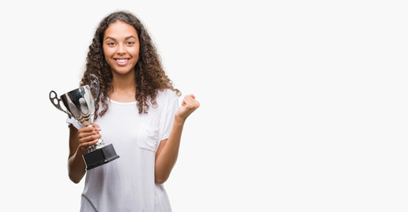 Wall Mural - Young hispanic woman holding trophy screaming proud and celebrating victory and success very excited, cheering emotion