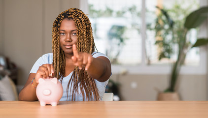 Wall Mural - African american woman saving money with piggy bank pointing with finger to the camera and to you, hand sign, positive and confident gesture from the front